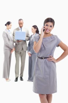Businesswoman on the phone with a hand on her hip, her legs crossing and her head tilted with co-workers in the background watching a laptop screen