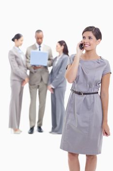 Businesswoman smiling on the phone with one arm along her body and co-workers in the background looking a laptop screen