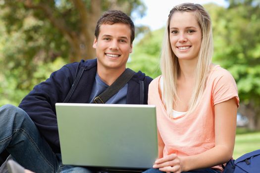 Portrait of two students laughing while sitting with a laptop in a park
