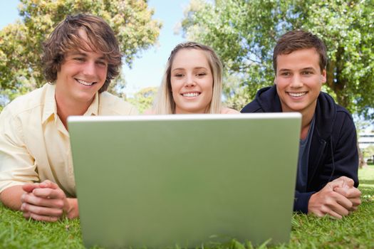 Three smiling students in a park lying while using a laptop