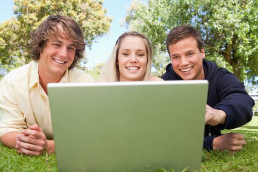 Portrait of three happy students in a park lying while using a laptop