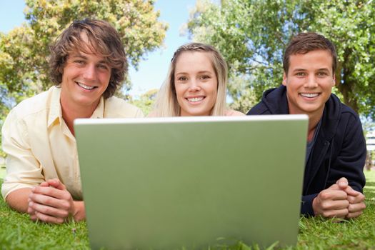 Close-up of three happy students in a park lying while using a laptop