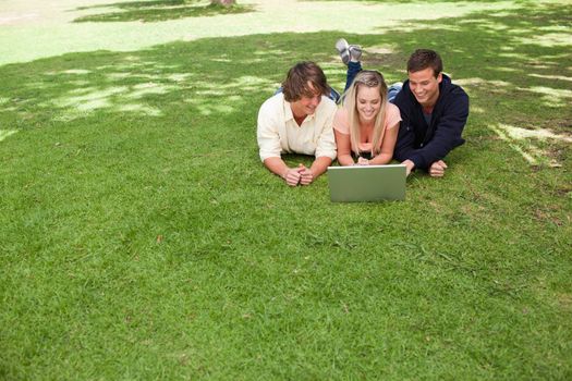 Three smiling young people in a park lying while using a laptop