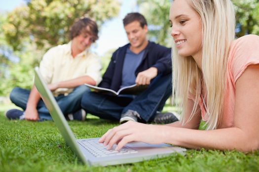Girl using a laptop while lying in a park with friends in background