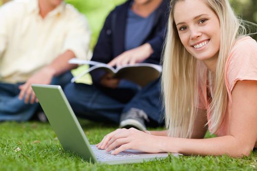 Portrait of a smiling girl using a laptop while lying in a park with friends in background