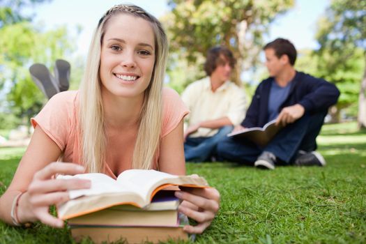 Smiling a girl lying in front of her books in a park with friends in background