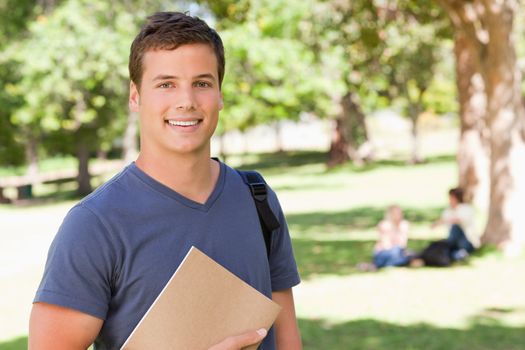 Portrait of a student smiling while holding a textbook in a park with friends in background