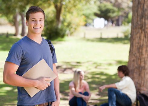 Portrait of a smiling student holding a textbook in a park with friends in background