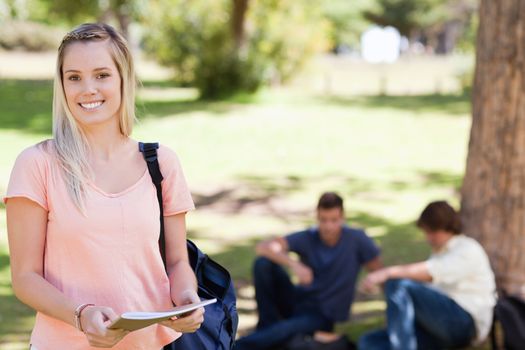 Portrait of a female student smiling while holding a textbook in a park with friends in background