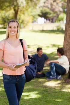 Portrait of a smiling female holding a textbook in a park with friends in background