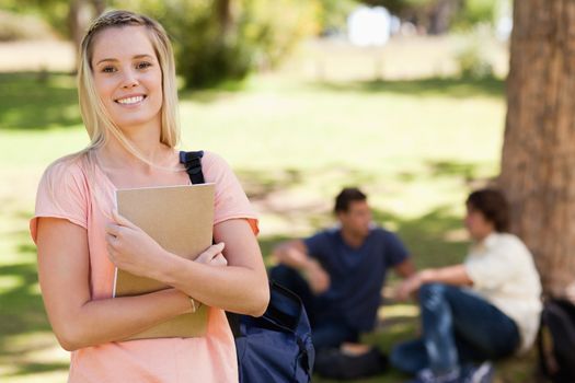 Portrait of a pretty girl smiling while holding a textbook in a park with friends in background