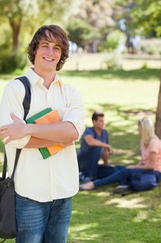 Smiling young man posing with textbook in a park with friends in background