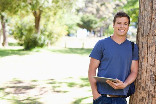 Portrait a student leaning against a tree with a touch pad in a park