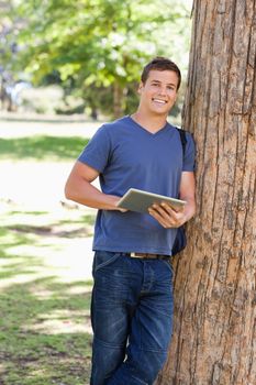 Portrait of a student leaning against a tree while using a touch pad in a park