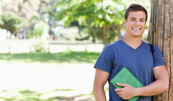 Portrait of a smiling muscled student holding a textbook in a park