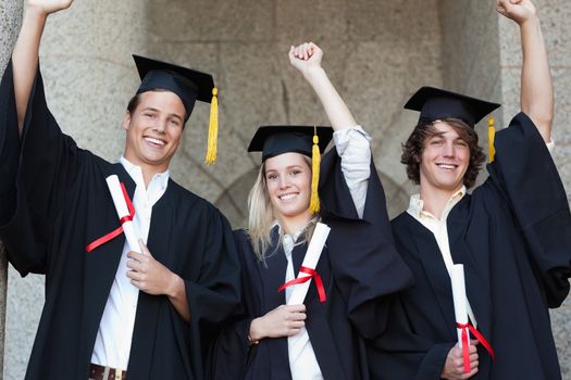 Graduates holding their diploma while raising arm with university in backgroung