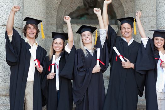 Smiling graduates posing while raising arms in front of the university
