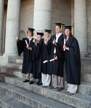 Laughing graduates posing the thumb-up in front of the university