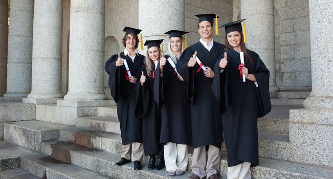 Five happy graduates posing the thumb-up in front of the university