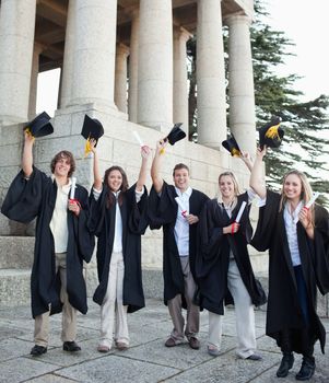 Five happy grad students raising their hats in front of the university
