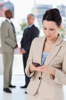Young serious businesswoman sending a text while executives are talking behind her