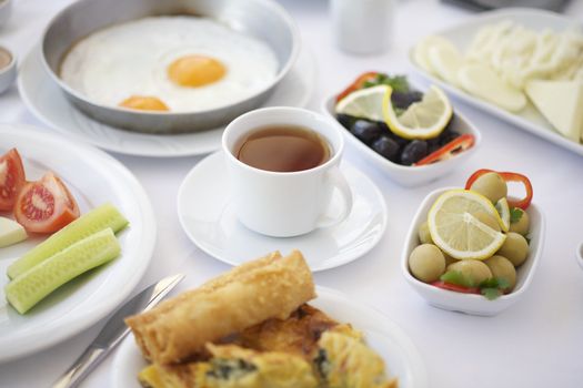 A cup of tea on breakfast table with tomatoes, cucumber, cheeses and olives