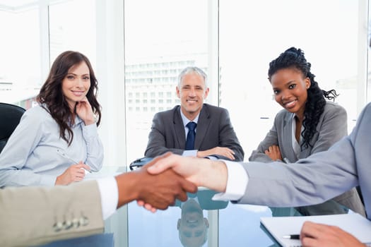 Three smiling colleagues looking at two executives shaking hands