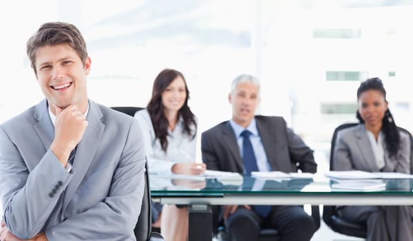 Young smiling businessman sitting in front of his team with his hand on his chin