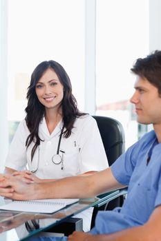 Enthusiastic female doctor sitting at the desk while looking at the camera near a co-worker