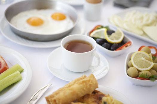 A cup of tea on breakfast table with tomatoes, cucumber, cheeses and olives