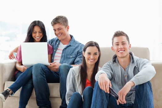 A man and woman sitting on the ground looking at the camera as another man and woman look at a laptop while sitting on the couch