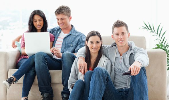 A smiling couple on the ground with a tv remote while another group sit on the couch together with a laptop