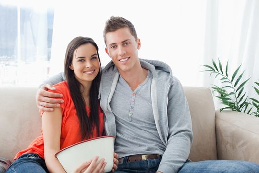 A couple with a bowl wit together on the couch as they look into the camera