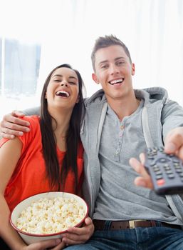 A couple sitting on the couch laughing as they have popcorn and look into the camera