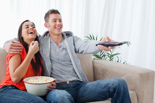 A laughing couple with popcorn as they change the tv with a remote and sit on the couch