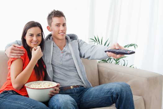 A smiling couple looking at the camera as they eat popcorn and use the tv remote