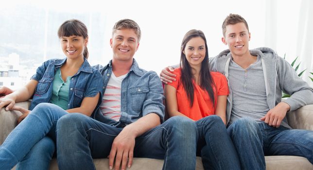 Two smiling couples sit on the couch beside one another as they look forward into the camera