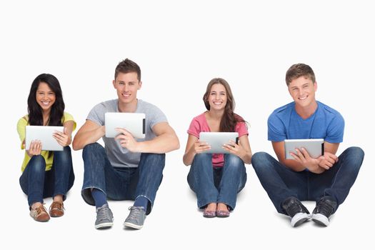 A smiling group of people sitting on the ground beside each other while using tablets and looking at the camera