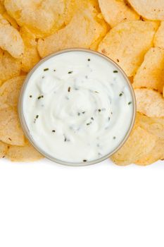 Close up of a bowl of white dip with herbs surrounded by nachos against a white background