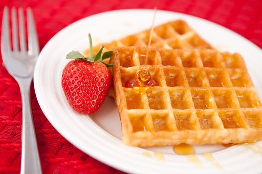 Waffles and syrup and strawberry together in a white plate om a red napkin