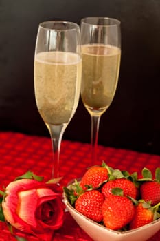Top glasses of champagne with strawberries in a bowl and a rose on a red tablecloth against a black background