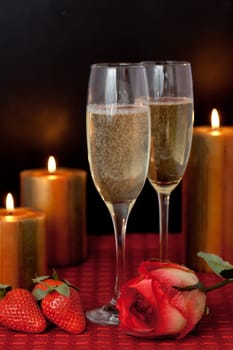Glasses of champagne with strawberries and a rose beside candles on a red tablecloth against a black background