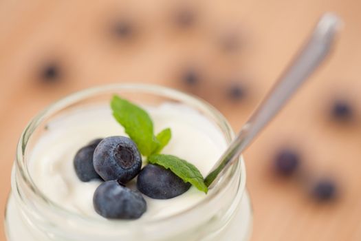 Close up of a pot of yoghurt with four leaves and blueberries and blurred blueberries in the background
