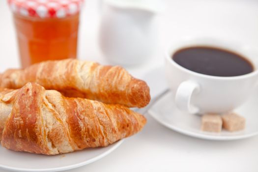 Croissants and a cup of coffee on white plates with sugar milk and a pot of jam against a white background