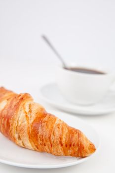 Croissant and a cup of coffee with a spoon on white plates against a white background