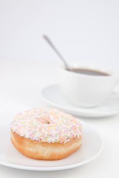 Doughnut with multi coloured icing sugar and a cup of coffee with a spoon on white plates against a white background