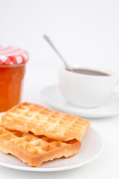 Waffles and cup of coffee with a spoon on plates and a pot of jam against a white background