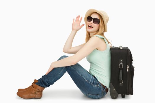Woman raising her hand while sitting next to a suitcase against white background