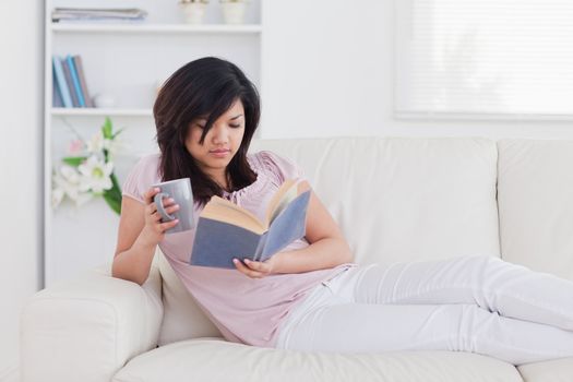 Woman lying on a sofa while holding a mug and a book in a living room