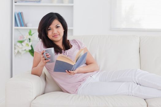 Woman lying on a couch while holding a mug and a book in a living room